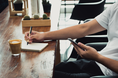 close up casual asian man use mobile phone and writing on notebook on wood table in coffee shop,Modern online working Digital lifestyle concept.influencer reviewの写真素材