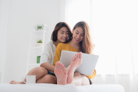 Asian lesbian lgbt couple sitting on bed hug and using laptop computer while drinking hot coffee cup and near white window sunlight in bedroom with happiness moment together,love wins conceptの写真素材