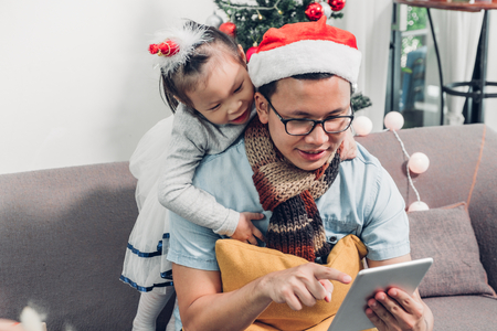 Father and daughter sitting on sofa  and select christmas present and shopping from online store by tablet at xmas party,holiday gift giving festiveの写真素材