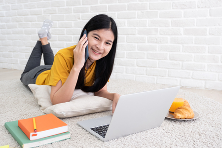 Asian female talk on mobile and use laptop computer to work at home.woman lay down on carpet at brick wall.working online lifestyleの写真素材