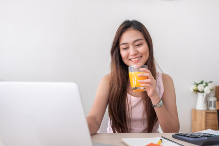 Asian female freelancer drink orange juice and and working on laptop at home.Work at home concept.work from home conceptの写真素材