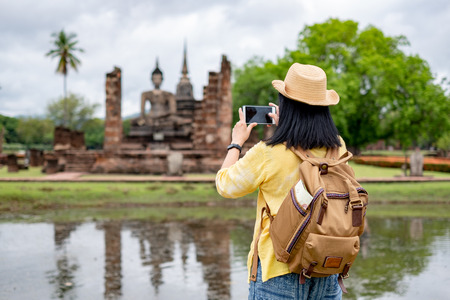 Asian tourist woman use mobile take a photo of ancient of pagoda temple thai architecture at Sukhothai Historical Park,Thailand. Female traveler in casual thai cloths style visiting city conceptの写真素材