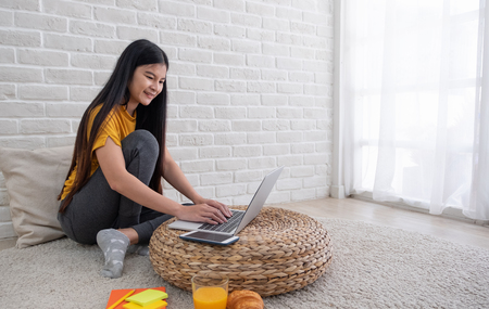 Asian female sit with knees up on floor using laptop on wicker stand near window in living room at home.Work at home concept.work from home.relax lifestyleの写真素材