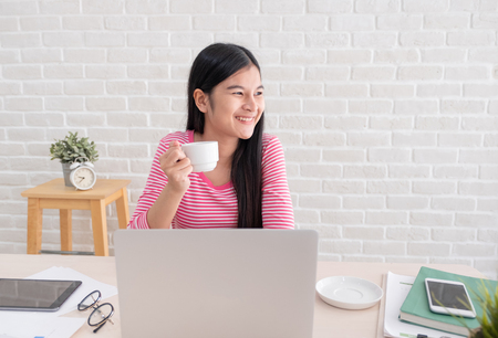 Asian female freelancer smile and drink coffee with relax emotion and looking out of window  take breake from laptop work on table at brick wall at home.working online lifestyleの写真素材