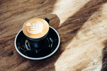 Close up hot black coffee cup with tree heart shape latte art milk foam on wood table near window with hard shadow sunlight at cafe restaurant.の写真素材