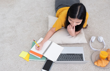 Top view asian woman lying on carpet and writing on notebook and learning on laptop with relax emotion at home,work at home conceptの写真素材