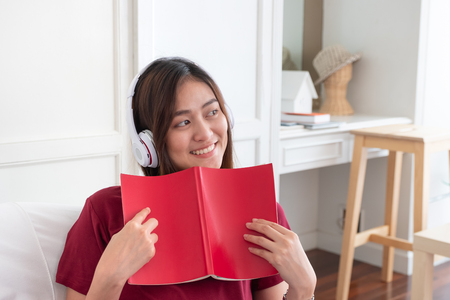 Asian woman listening music on headphone and reading book with relax pose at couch in living room at home.leisure lifestyleの写真素材