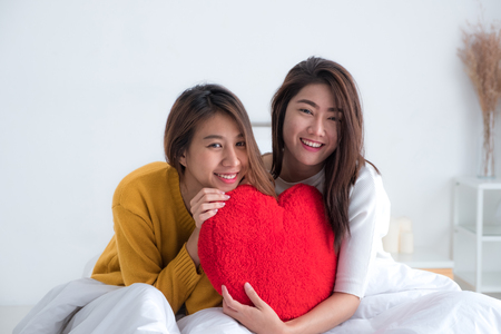 Asia lesbian lgbt couple holding red heart pillow together and sitting on sofa in living room.Love wins conceptの写真素材
