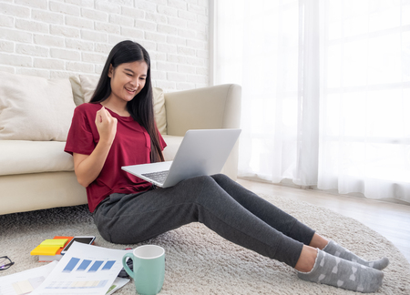 Asian woman freelancer hand up and smile for success working on laptop on sofa at home.work at home concept.yeah emotionの写真素材