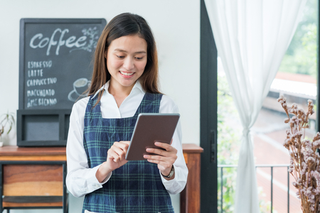 Asian female barista wear blue apron hold tablet computer coffee menu with smiling face in cafe waiting order from customer ,Coffee business owner conceptの写真素材
