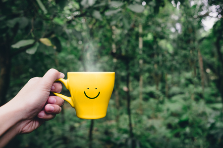 Hand holding hot yellow coffee cup with hand drawn smile face on cup at tropical nature forest,Leisure lifestyleの写真素材