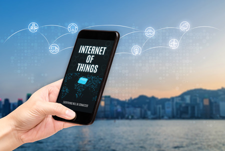 Hand holding mobile with Internet of things (iot) word on screen with icon feature glowing in air with blur city building background,Digital Lifestyle conceptの写真素材