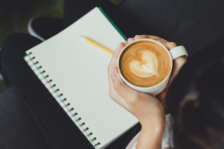 Top view woman hand holding hot cappuccino coffee cup with heart latte art with notebook and pencil in cafe shop,leisure activityの写真素材