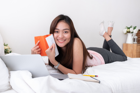 Asian woman wear pink vest lying on bed reading book with research job on laptop in bedroom.work at home.e-larning concept.study onlineの写真素材