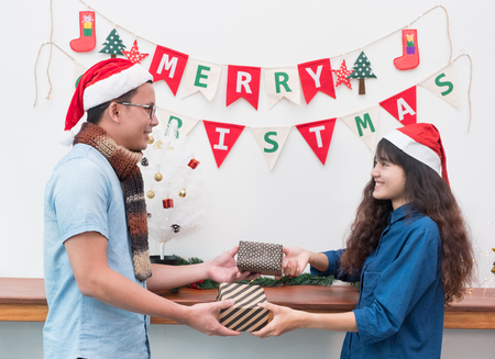 Asian lover couple wear santa hat in merry christmas party and exchange gift box each other with smiling face,Xmas gift giving,holiday celebration festive seasonalの写真素材