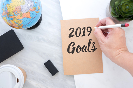 Hand holding pen writing 2019 Goals on brown paper with blue globe,blackboard,coffee cup on marble table.new yearâs resolutions conceptの写真素材