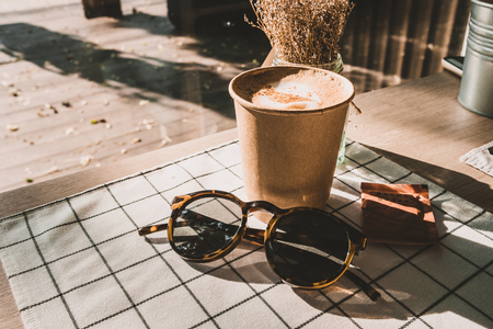 cappuccino in take away coffee cup and sunglasses on table cloth with dry flower on wood table with sunlight hard shadow at window in evening.food and drink lifestyle leisure conceptの写真素材