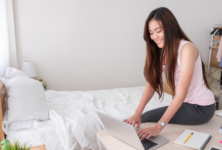Asian female freelancer wear vest shirt typing on laptop on table and thinking about job in bedroom at home.Work at home.doing paperwork from homeの写真素材
