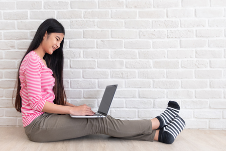 Asian woman in casual pink cloth use laptop and sitting on floor at white brick wall in house.mock up screen and copy space for adding text or designの写真素材