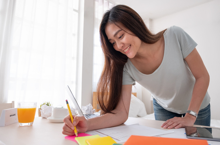 Asian female freelancer standing and writing on paperwork working on table in bedroom at home.Work at home concept.work from home conceptの写真素材