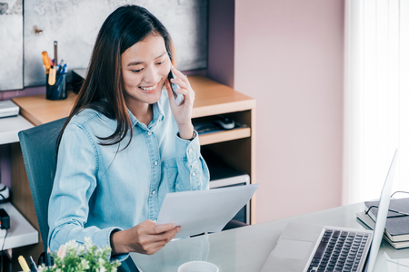 Asian businesswoman talking on mobile phone with customer and looking to work sheet paper in front of laptop computer at office,Office lifestyle conceptの写真素材