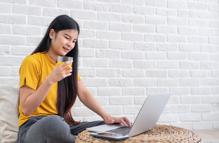 Asian female sit on floor using laptop on wicker stand and drink orange juice in living room at home with relax emotion.Work at home concept.work from home.relax lifestyleの写真素材
