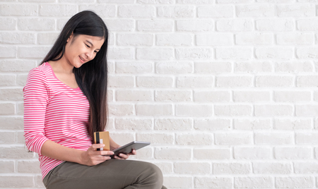 Asian woman in casual cloth holding credit card and using to online shopping with tablet at white brick wall.Digital technology lifestyle.Banner with copy space for display text or designの写真素材