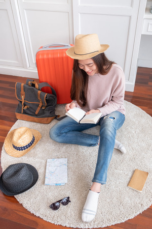Young asian woman traveler prepare things into orange suitcase for holiday vacation at home.backpacker travel conceptの写真素材