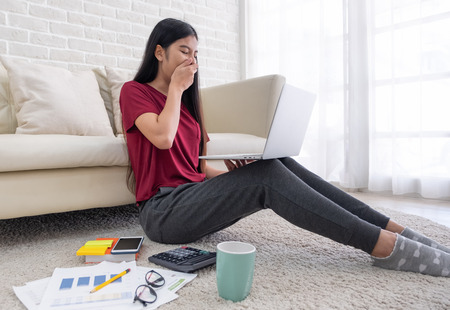 Asian woman freelance yawn while working on laptop with paperwork sitting one floor and leaning sofa at home,work at home conceptの写真素材