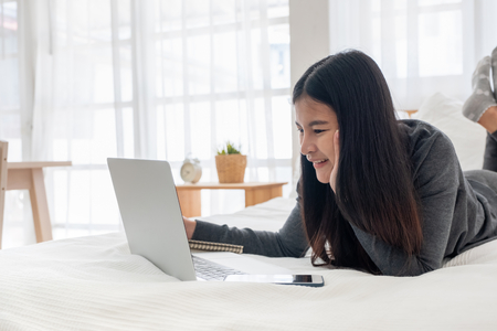 Asian woman laying on bed doing paperwork on laptop in bedroom.work at home.e-larning concept.study onlineの写真素材