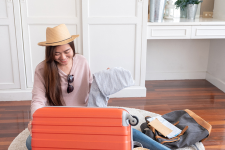 Young asian woman traveler prepare things into orange suitcase for holiday vacation at home.backpacker travel conceptの写真素材