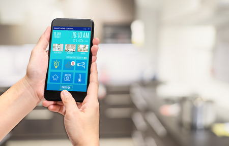 Hand holding mobile use smart home control mobile app at blur bokeh kitchen background,smart living concept,digital lifestyleの写真素材