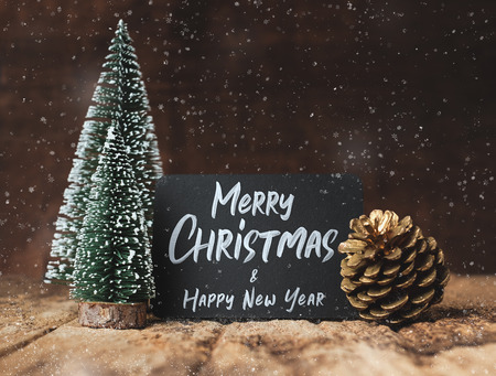 Merry Christmas and happy new year on blackboard with xmas tree and gold pine cone and snow falling on grunge wood table and dark brown wooden wall.winter holiday greeting cardの写真素材