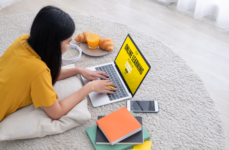 Asian female student  log on to e-learing website on laptop computer at home.woman lying down on carpet.learning online lifestyleの写真素材