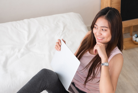 Asian female freelancer smile and holding laptop with rest leg on table in bedroom at home.Work at home concept.work from home conceptの写真素材