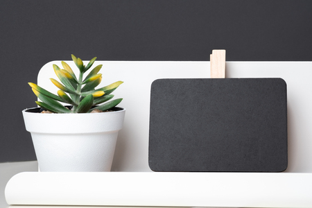 Black clip blackboard on modern pencil box and green plant in white pot on table and dark grey wall in office desk,mock up space for display of design or textの写真素材