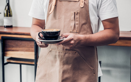 asian male barista wear apron holding hot coffee cup served to customer with smiling face at bar counter,Cafe restaurant service concept.waitress workingの写真素材