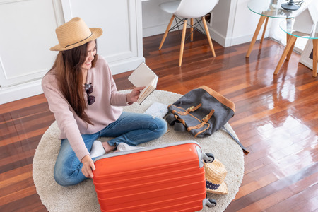 Young asian woman traveler prepare things into orange suitcase for holiday vacation at home.backpacker travel conceptの写真素材
