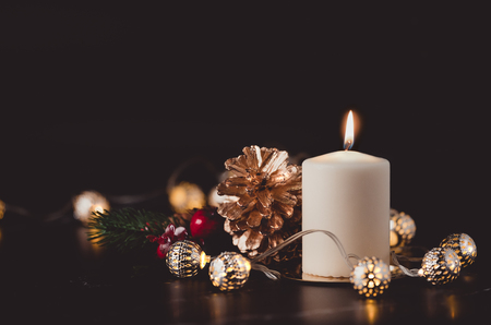 Christmas white candle with golden pine cone,mistletoe and light string glowing on black marble table with dark background.holiday greeting cardの写真素材