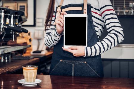 Asian woman barista wear jean apron holding blank screen tablet to customer at bar counter with smile emotion,Cafe restaurant service concept,Owner small business.mock up device for display textの写真素材