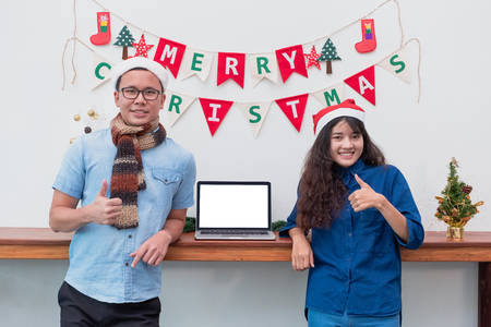 smiling Lover asian couple thumbs up and standing at Christmas garland on wall with with laptop,Mock up template blank screen computer for display of designの写真素材
