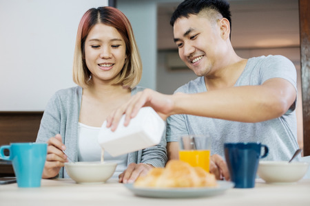 happy asian couple in pajamas sitting at table in kitchen at home in morning and having cereal breakfast together.husband pour milk into wife bowl.の写真素材