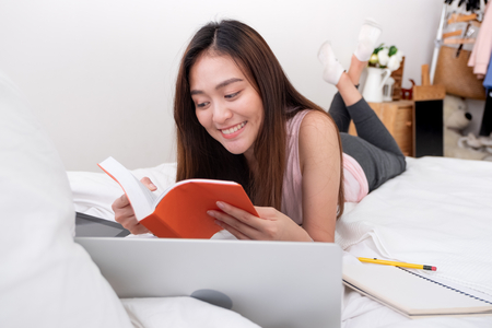 Asian woman wear pink vest lying on bed reading book with research job on laptop in bedroom.work at home.e-larning concept.study onlineの写真素材