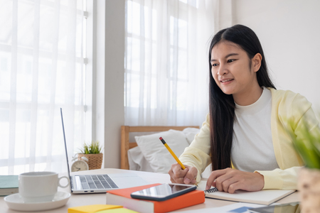 Asian female freelancer writing paperwork while working with laptop on table in bedroom at home.Work at home concept.work from home conceptの写真素材