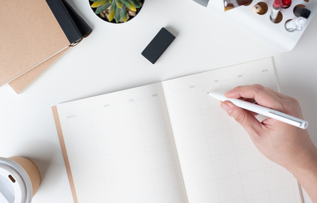 top view of hand writing on open calendar planner for business resolution with modern office stationery and take away coffee cup on white desk in office.mock up space of organizer table notebookの写真素材