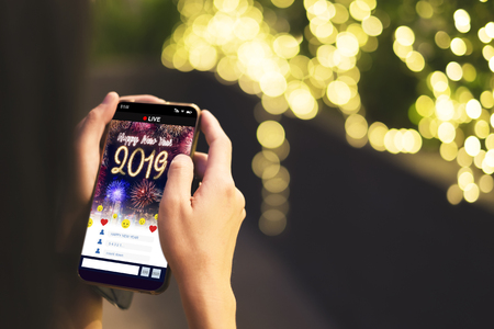 Woman Hand holding mobile see live streaming countdown to Happy new year 2019 with fireworks over cityscape on screen with light bokeh background,Digital social media lifestyleの写真素材