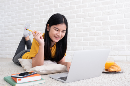 Asian female freelancer working on laptop computer at home.woman lying down on carpet at brick wall.working online lifestyleの写真素材