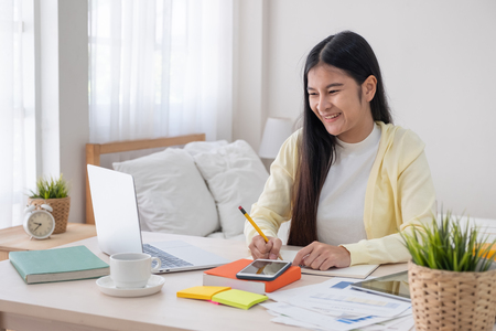 Asian female freelancer reading report on laptop and jot down note on table in bedroom at home.Work at home concept.work from home.relax lifestyleの写真素材