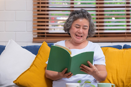 Asian senior woman sitting and reading book at sofa at home in relax emoitonの写真素材