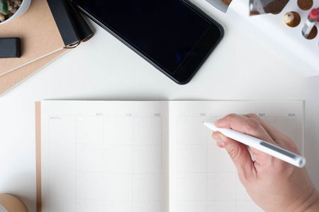 top view of hand writing on open calendar planner for business resolution with modern office stationery and take away coffee cup on white desk in office.mock up space of organizer table notebookの写真素材